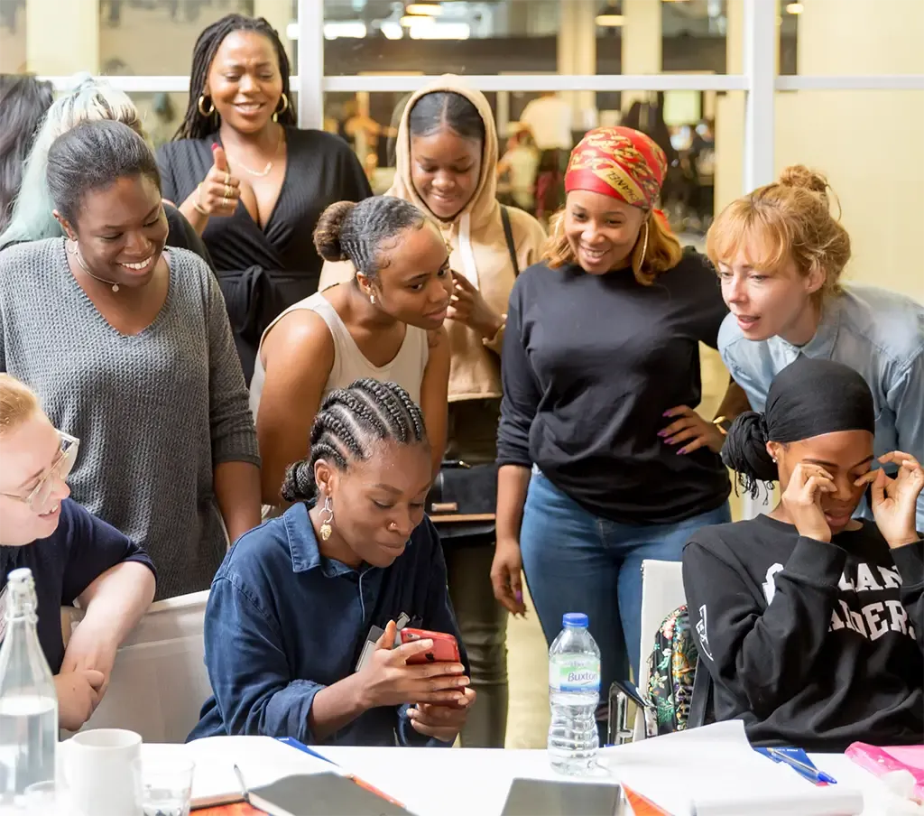 Women gathered around a table, looking at a phone held by a seated woman.