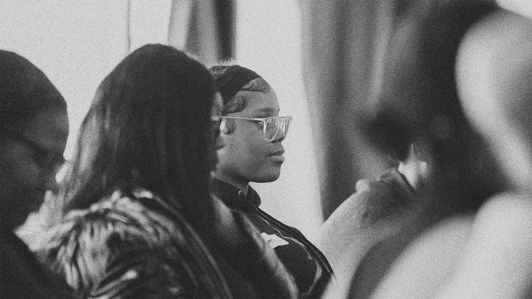 Women seated together, with a woman in clear-framed glasses in focus.