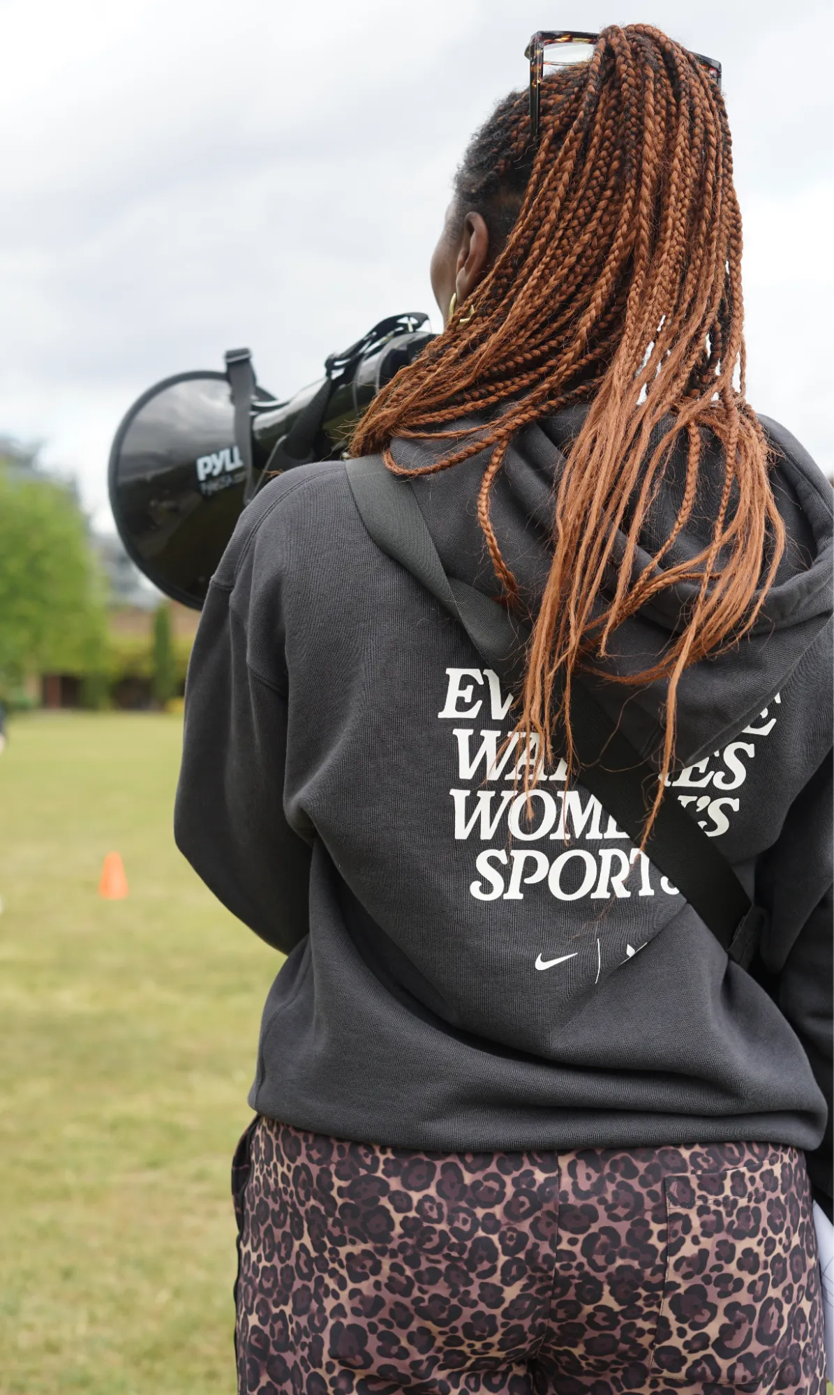 A person seen from behind holding a megaphone on a grass playing field, with a sports cone nearby.