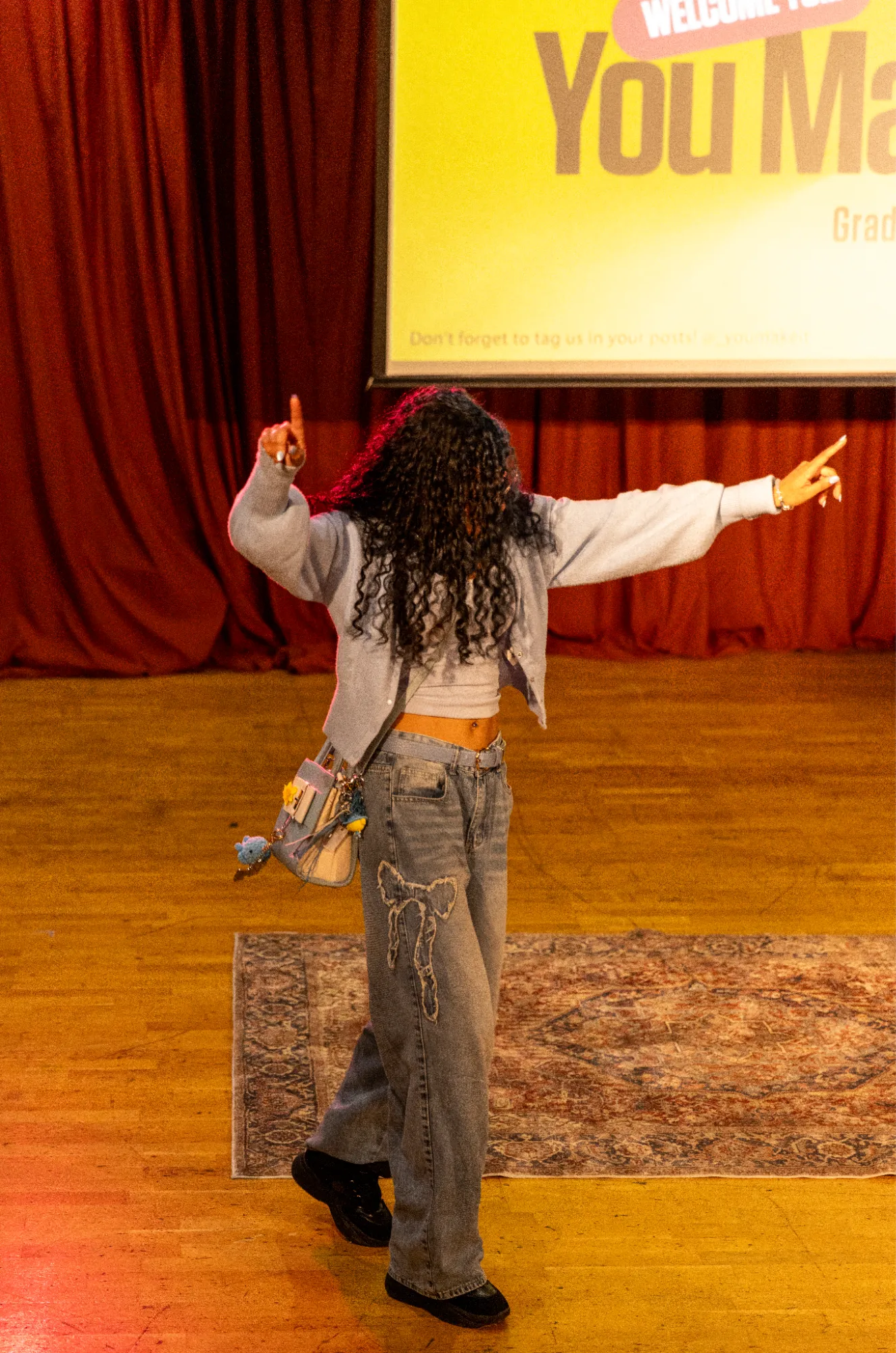 A person dancing with arms outstretched in front of stage curtains and a You Make It projection.