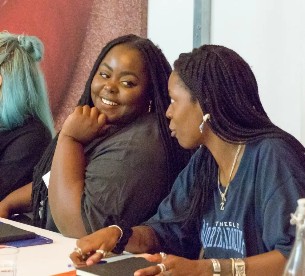Two women seated beside each other at a table, one smiling towards the other.